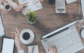 Four pairs of hands in business attire over a table strewn with laptops, calculaors, tablets, papers, and cofee mugs.