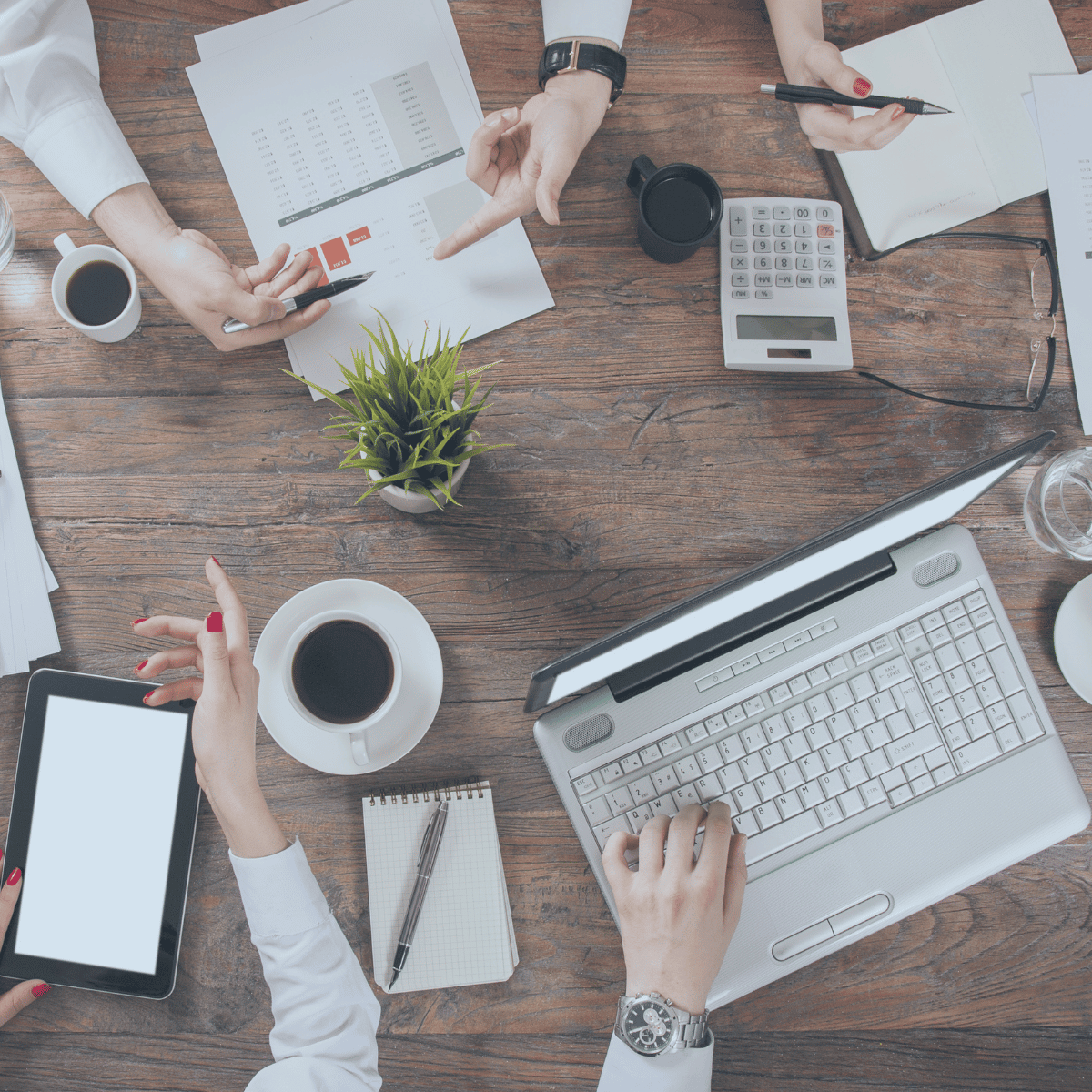 Four pairs of hands in business attire over a table strewn with laptops, calculaors, tablets, papers, and cofee mugs.
